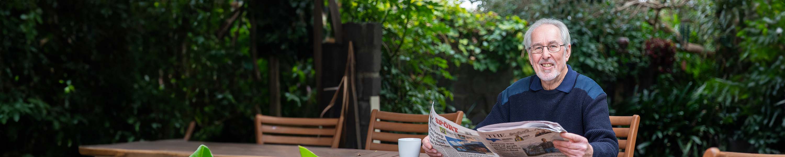 Older man reading newspaper in backyard, sitting at a table, surrounded by green plants