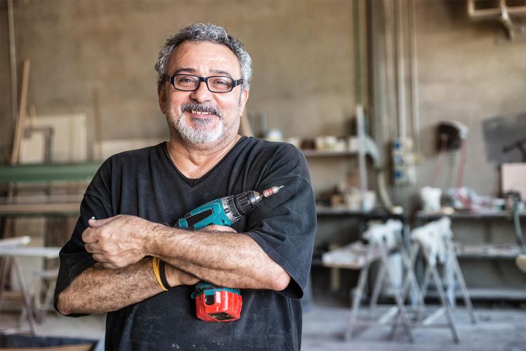 Older man in a workshop wearing a dark grey T-shirt, holding a power drill