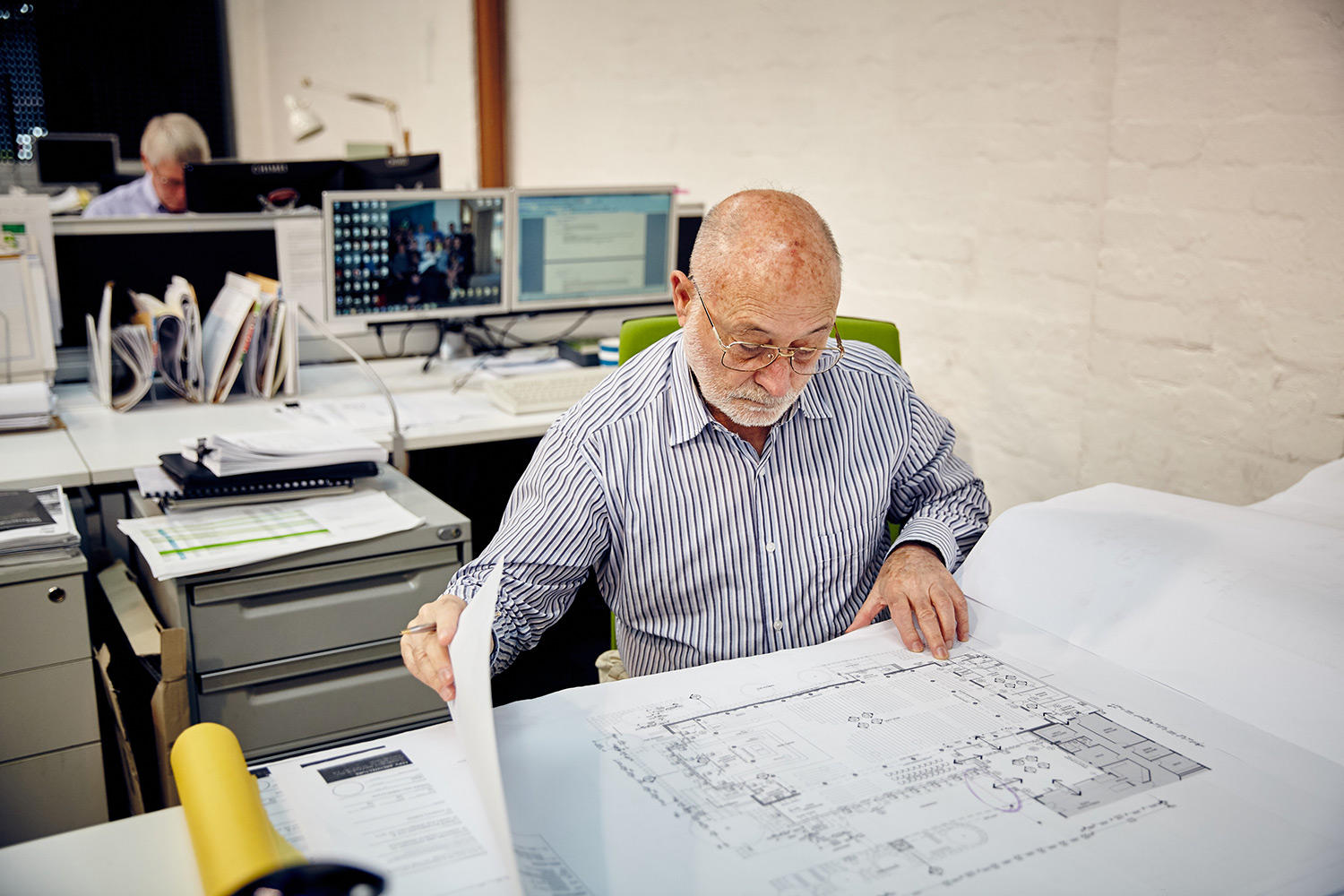 Older man in an office flipping through papers of structural drawings at desk